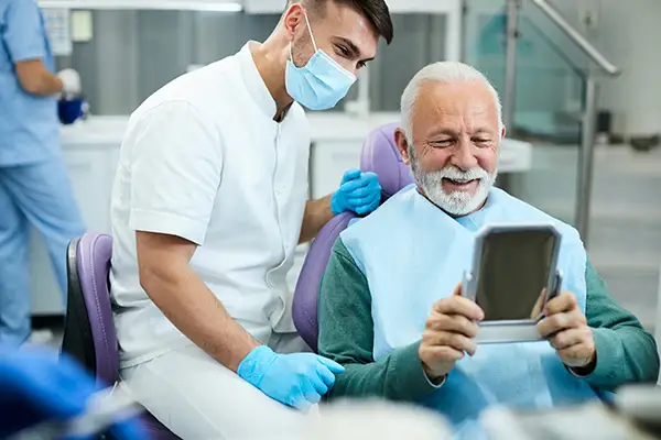 A happy senior patient looking at himself in a mirror with a dental assistant at Highridge Dental Care